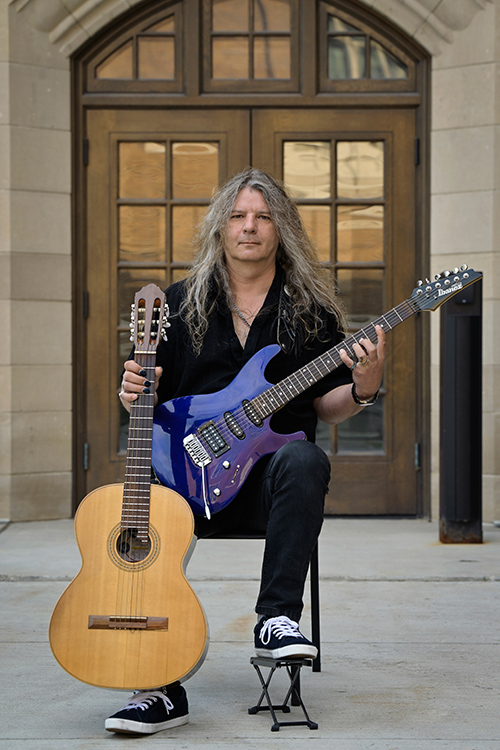 Photo of Corey holding an acoustic guitar and an electric guitar
