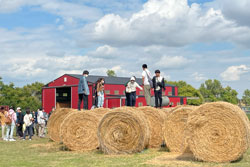 Students standing on hay bales in front of red barn on a sunny day