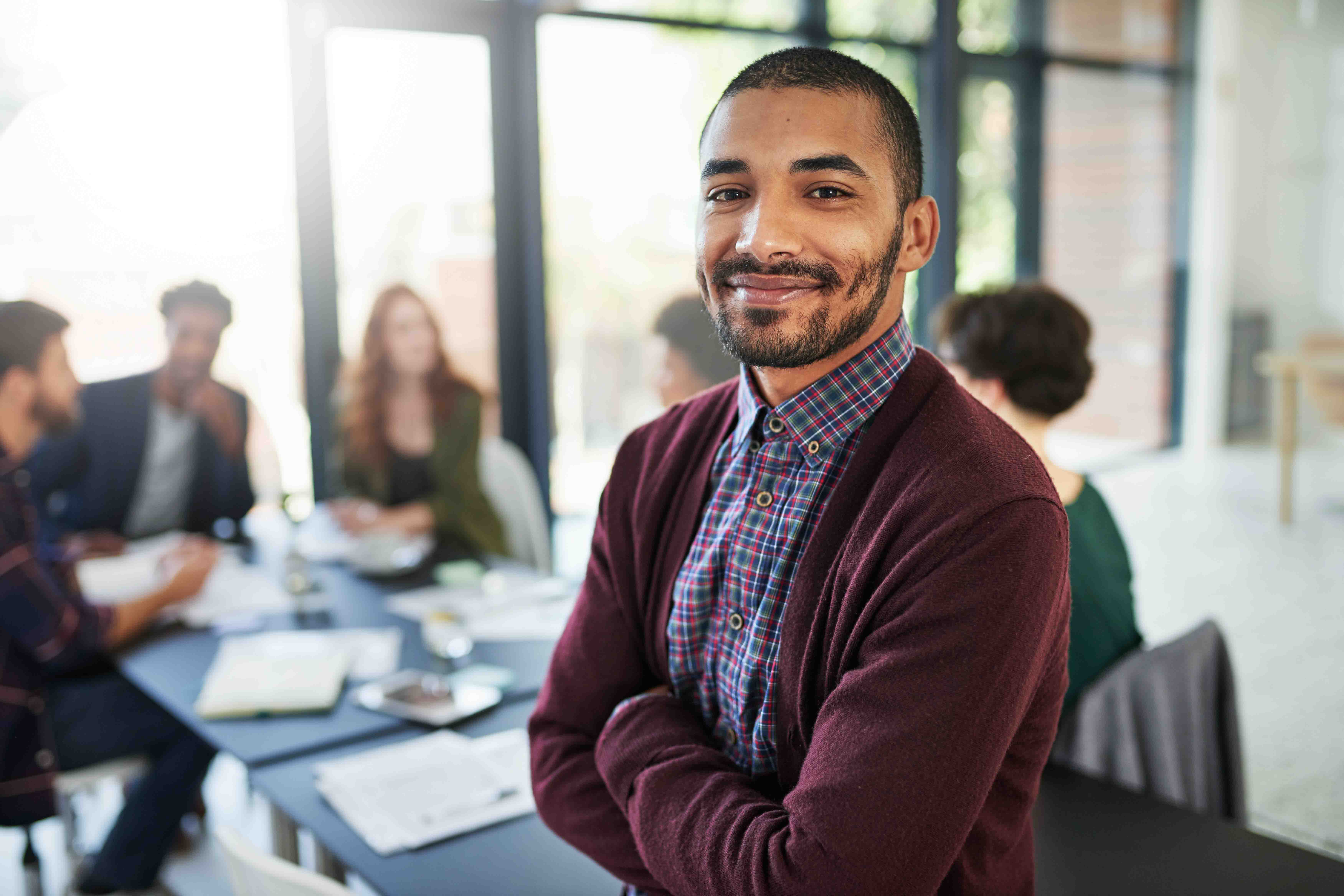 Mid-career man smiling & standing with arms crossed in front of board room table with other people working